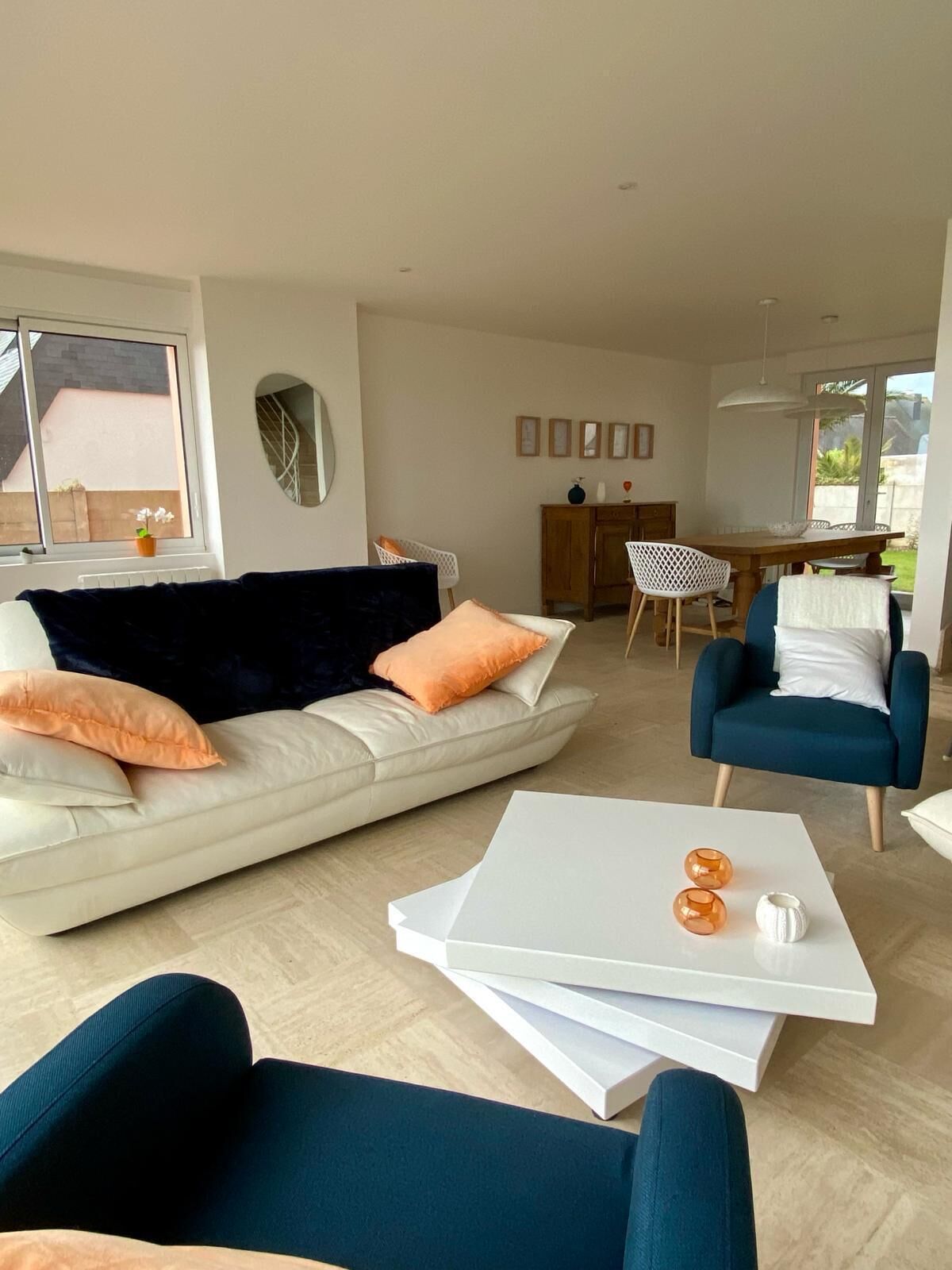 Un salon moderne avec un canapé blanc, des fauteuils bleus et une table basse blanche. On aperçoit à l’arrière-plan un coin repas avec une table et des chaises en bois.