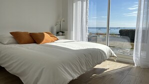 Una camera da letto con un letto grande, biancheria da letto bianca e vista sul mare attraverso tende leggere.