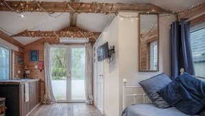 Ein gemütliches Blockhaus-Interieur mit Holzbalkendecke, einem Bett mit blauer Bettwäsche, einem an der Wand befestigten Fernseher und Blick ins Freie durch eine Tür.