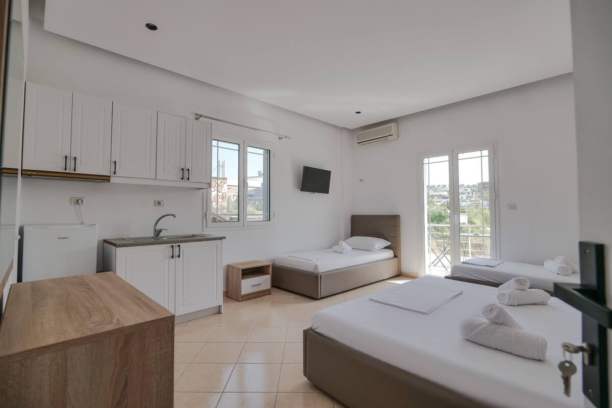 Habitación de hotel moderna con kitchenette, dos camas, un televisor y un balcón con vista a la ciudad.