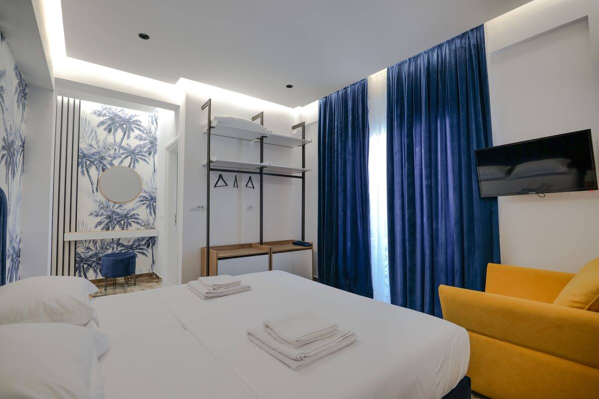 Una habitación de hotel con cama, una silla, un televisor, un estante con toallas y una ventana con cortinas.
