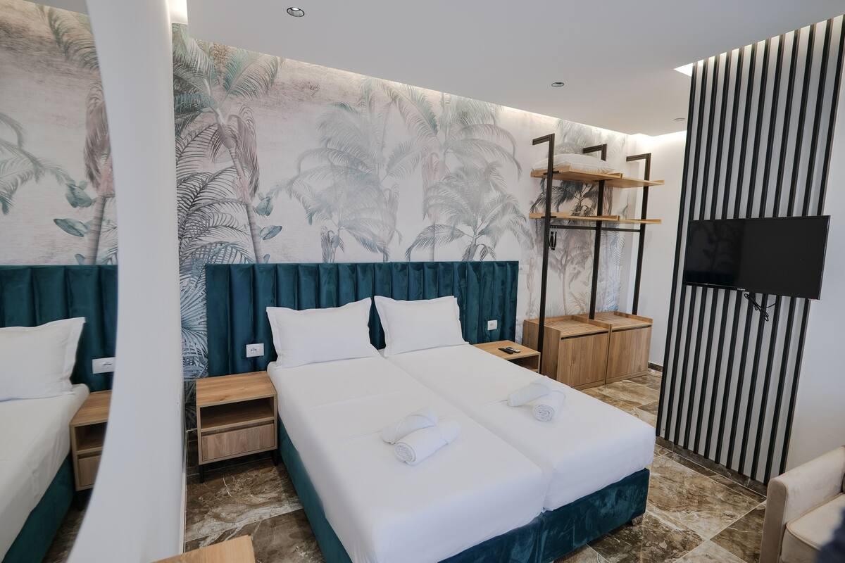 Habitación de hotel con una cama grande, dos mesitas de noche, un televisor y una pared con un mural tropical.
