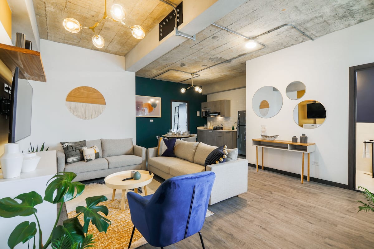 Un salon moderne comprenant un canapé, un fauteuil et une table basse, un coin cuisine avec des placards et des appareils électroménagers, ainsi qu’une table à manger avec des chaises.