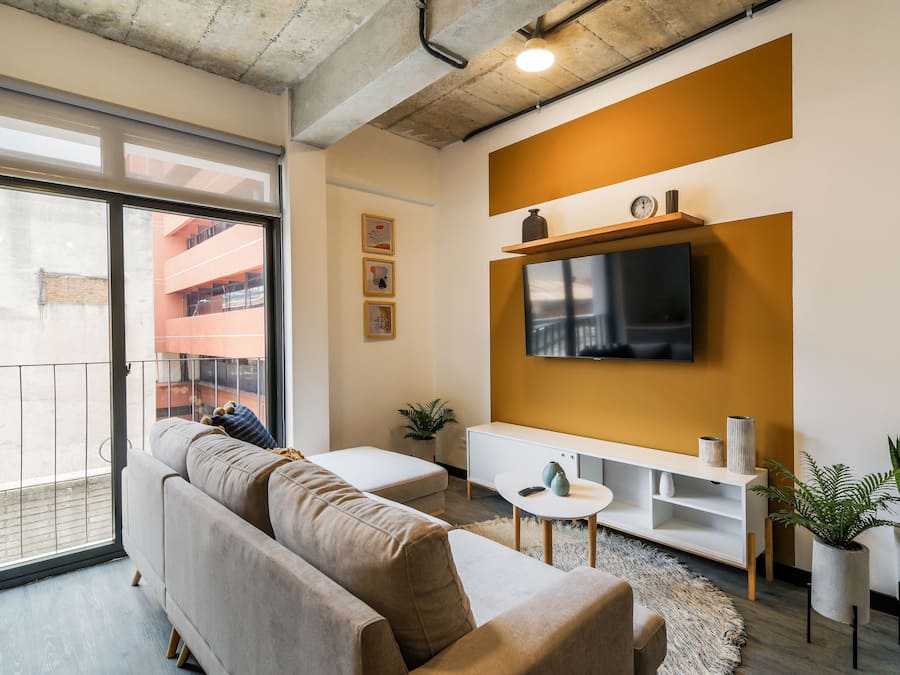 Ein modernes Wohnzimmer mit einer Couch, einem an der Wand montierten Fernseher, einem kleinen weißen Tisch und einem Blumentopf.