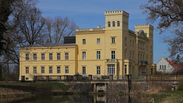 A large yellow building with multiple windows and a tower, situated next to a body of water. - Ehemalige Bibliothek: Küche Kamin Netflix Hofladen (Steinhöfel)