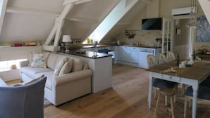 Ein geräumiges Loft mit hoher Decke, freiliegenden Balken, einer Küche mit weißen Schränken, einem Essbereich mit Holztisch und einer gemütlichen Sitzecke mit Sofa und Sesseln.