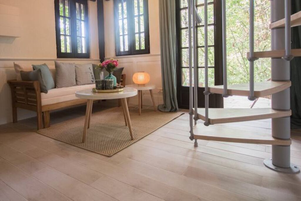 Une pièce avec un canapé en bois, une table ronde ornée d’un vase de fleurs, une petite lampe et un escalier en colimaçon.