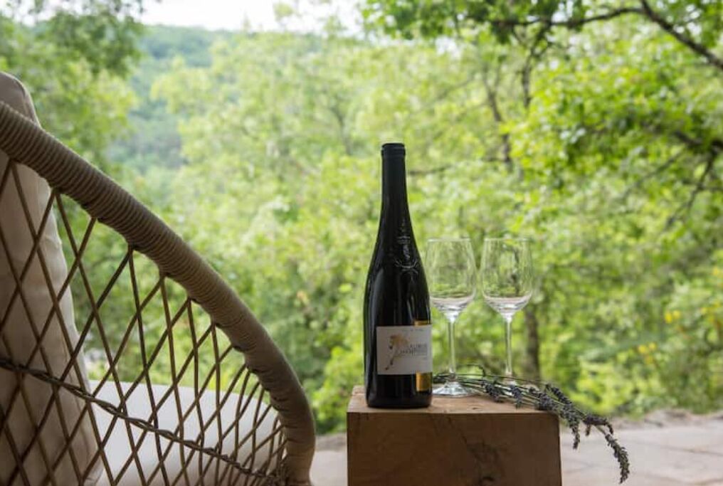 A wicker chair with a bottle of wine and two glasses on a wooden block, set against a lush green background.