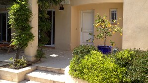 Un bâtiment avec un toit recouvert de tuiles, une porte et un petit arbre aux fruits jaunes. Devant la porte, on trouve un bac en béton dans lequel sont plantés des arbustes verts.