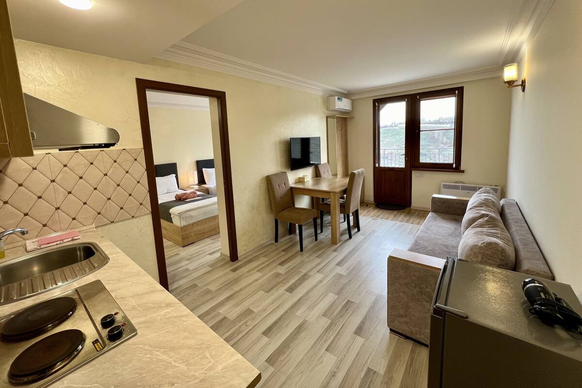 A compact hotel room with a kitchenette, a dining area, a sofa, and a TV.