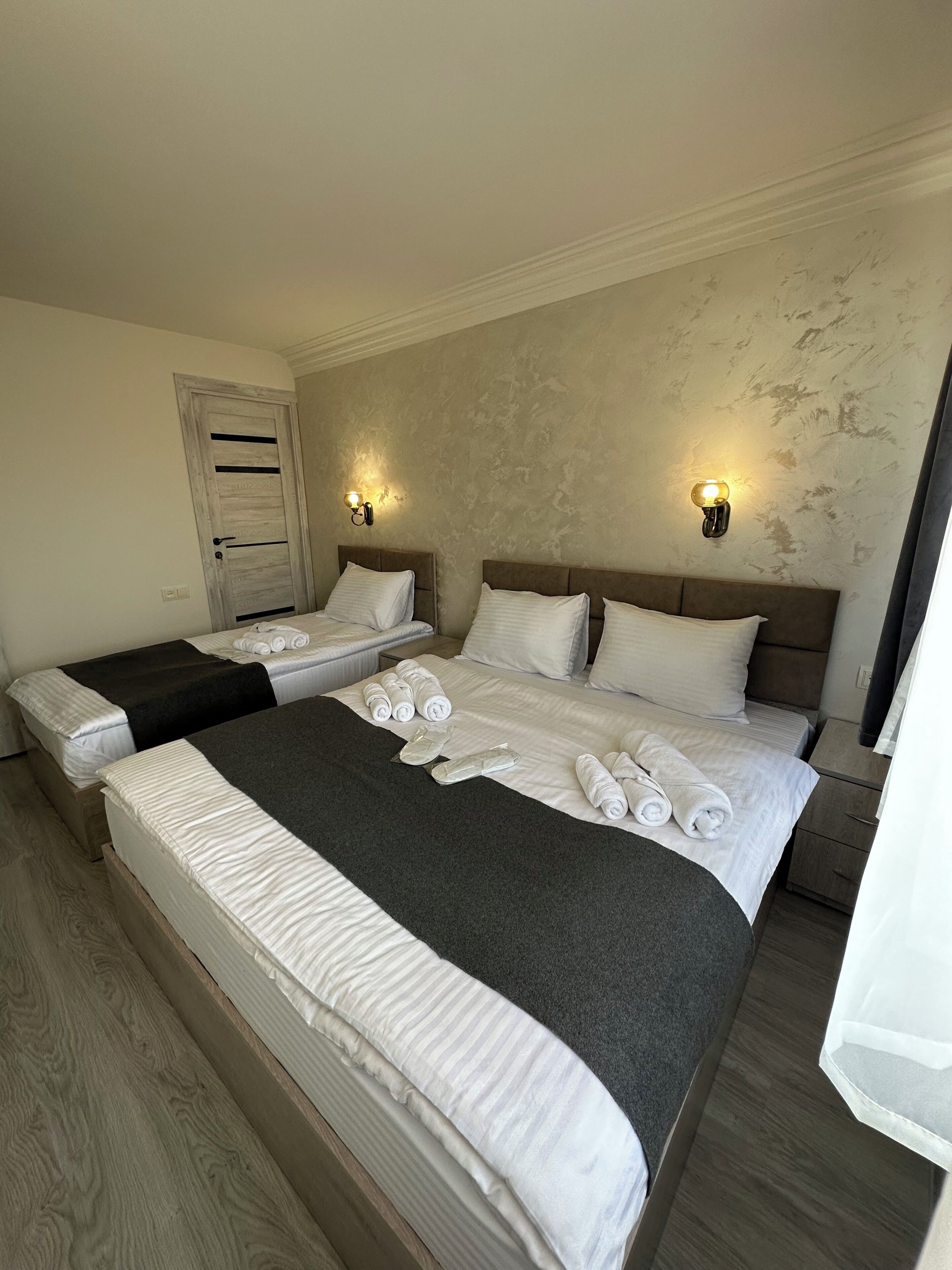 A hotel room with two beds, a large bed with a dark headboard, and a smaller bed with a light-colored headboard. There are towels neatly arranged on the beds, and two wall-mounted light fixtures.