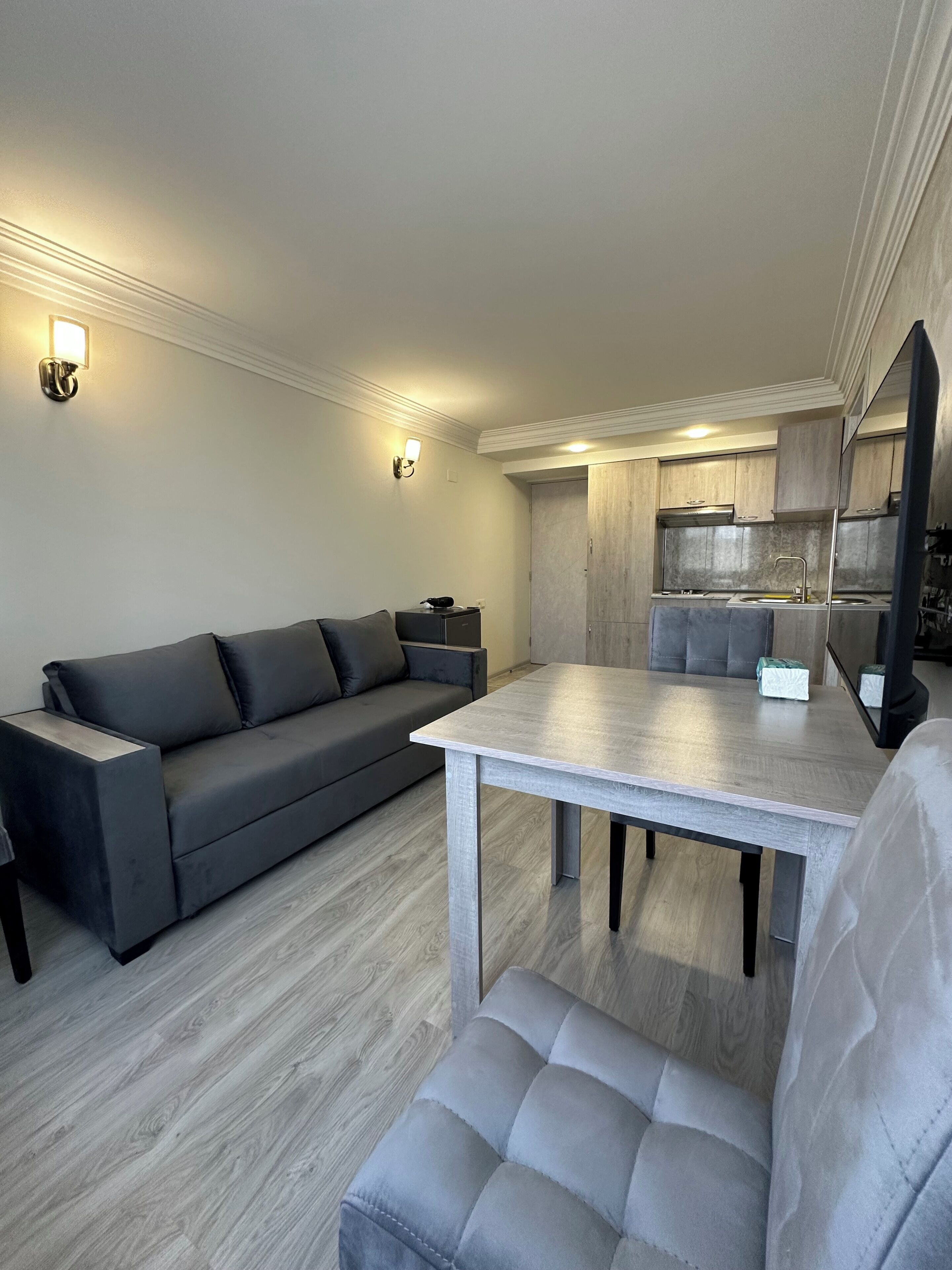 Una sala moderna con un sofá gris, una mesa de comedor de madera y una kitchenette al fondo.