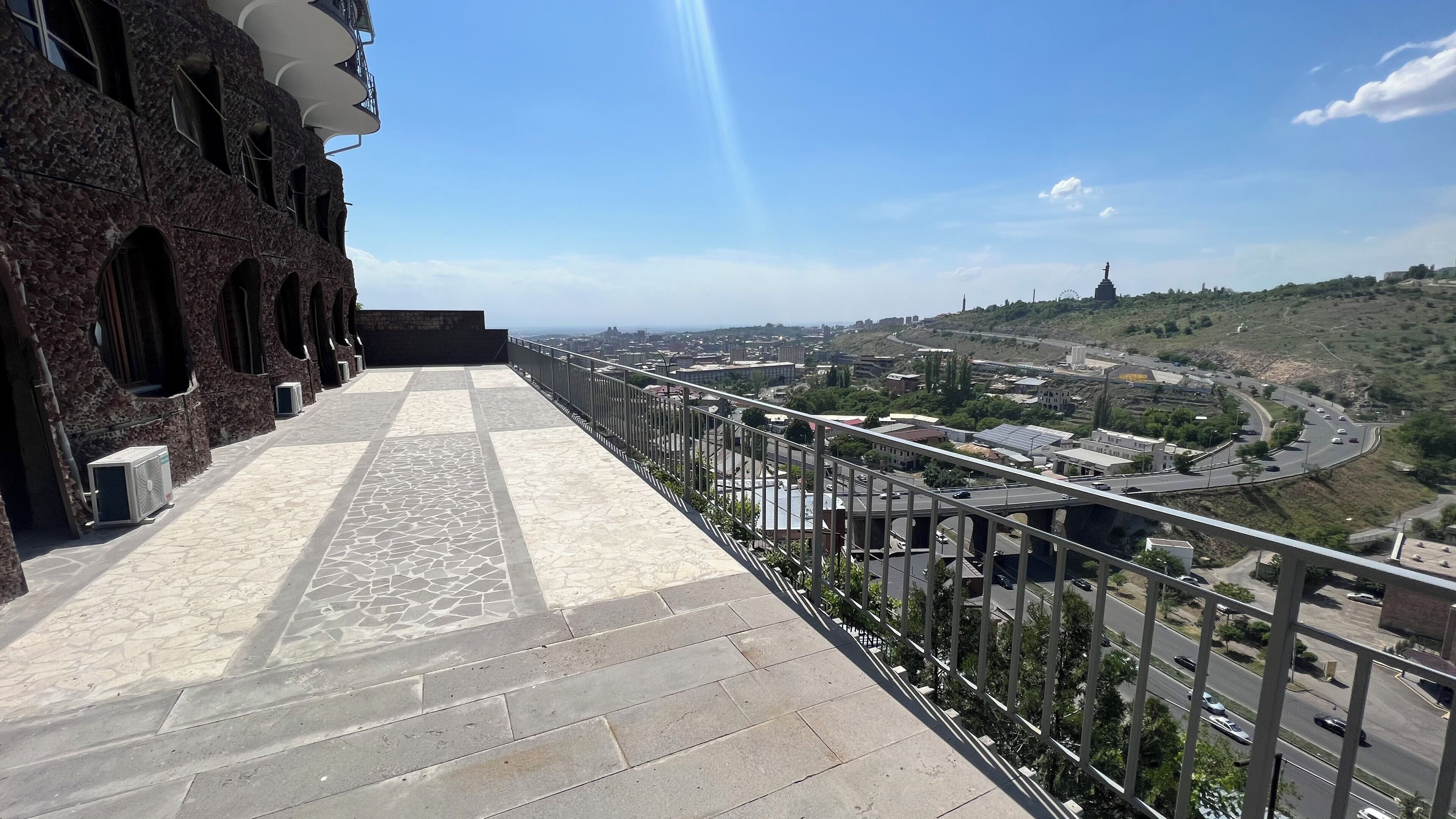 A balcony with a stone floor and a view of a cityscape with buildings and roads.