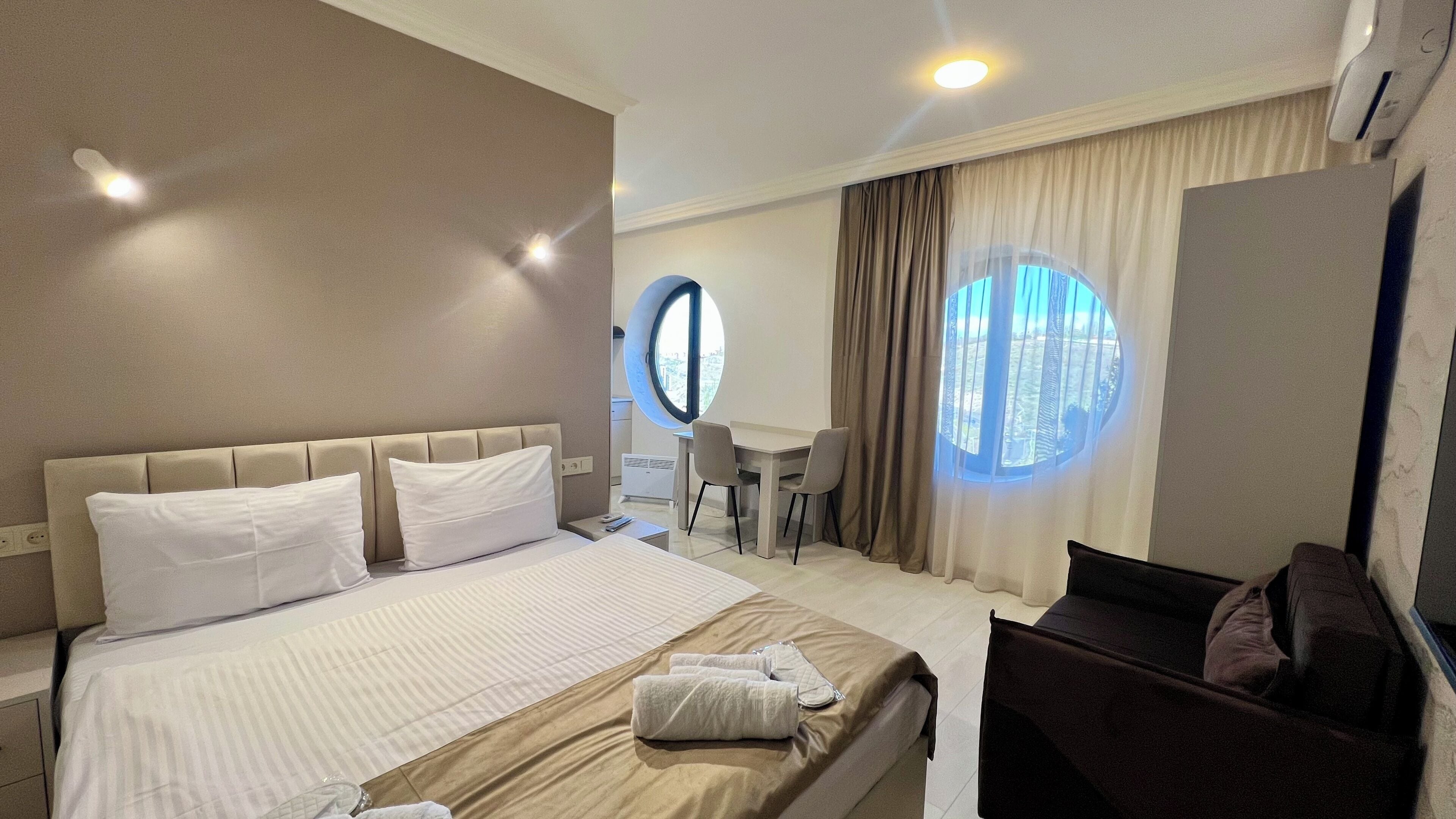 A hotel room with a bed, a desk, two chairs, a round window, and a view of the cityscape.
