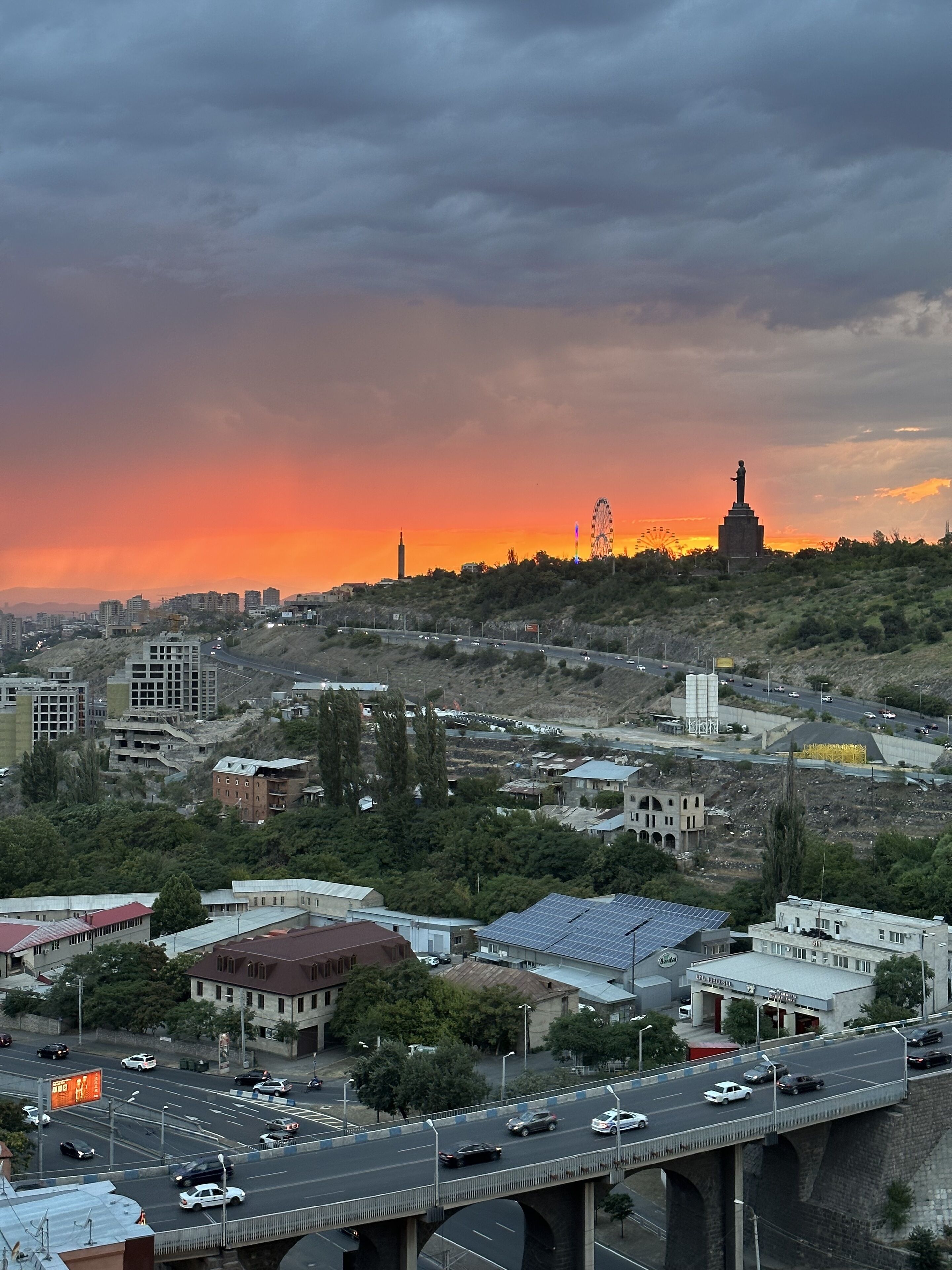 A cityscape with a prominent statue on a hill, a Ferris wheel, and a sunset.
