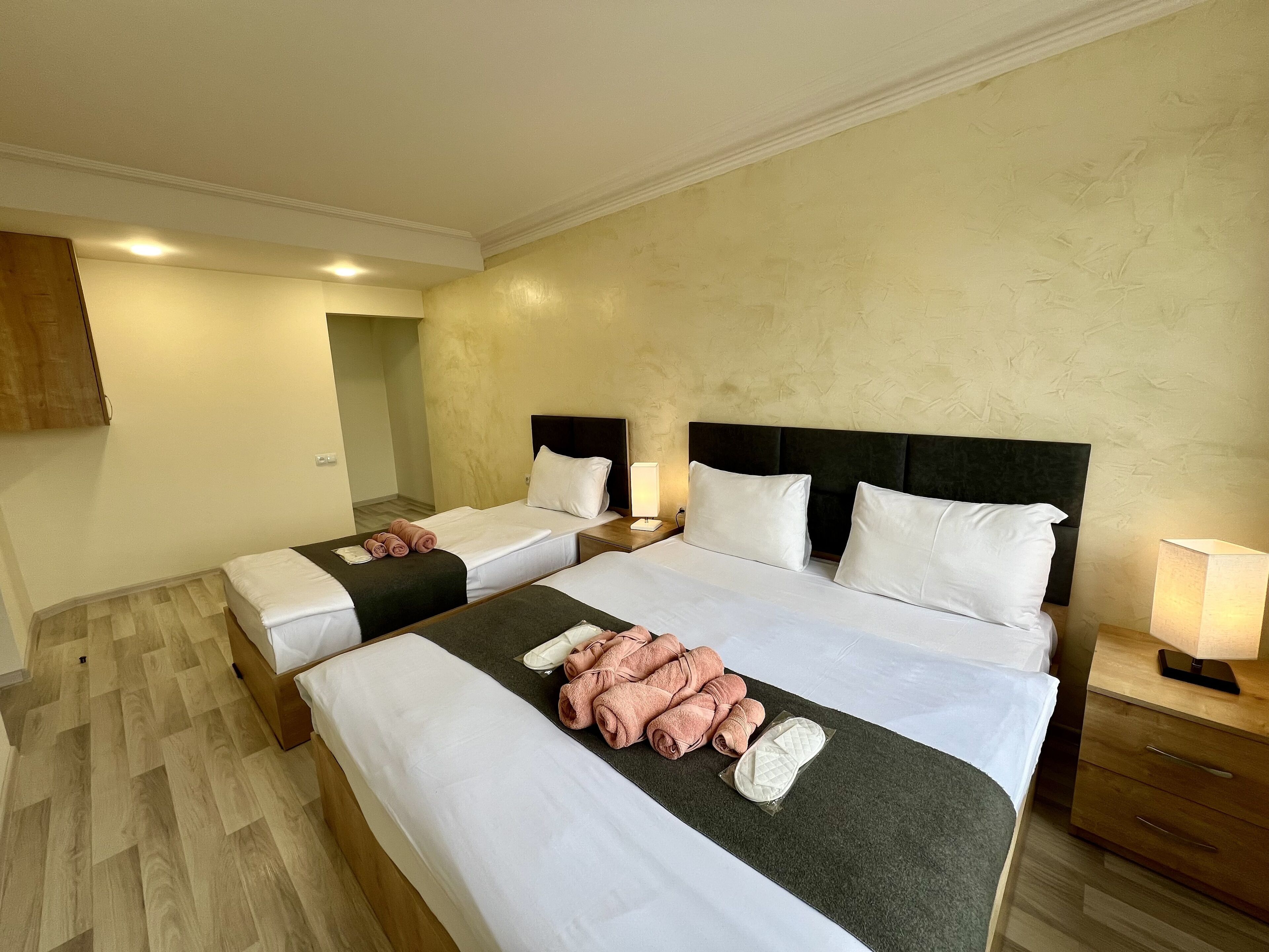 A hotel room with two beds, each with white linens and a dark headboard. There are towels and slippers on the beds, and a nightstand with a lamp.