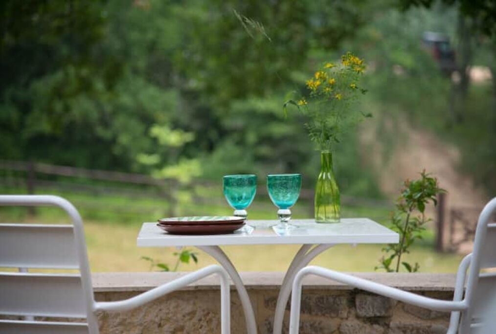 A table with two turquoise glasses and a green bottle, set against a stone wall with a view of a forested area.