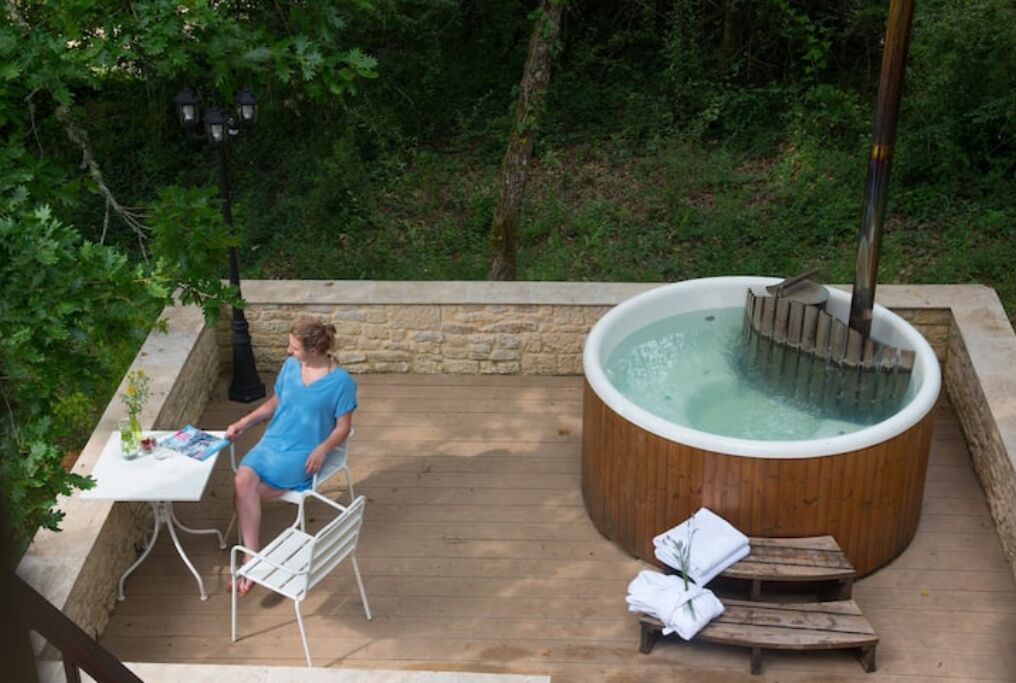 A wooden hot tub surrounded by a stone wall, with a table and chairs nearby.