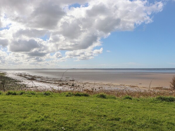 A beach with a grassy foreground and a wide expanse of water under a partly cloudy sky.