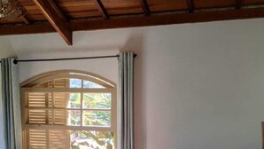 A bedroom with a wooden ceiling, a bed with patterned bedding, a window with shutters, and a view of greenery outside. - Canto dos Pássaros (Santo Antonio do Pinhal)