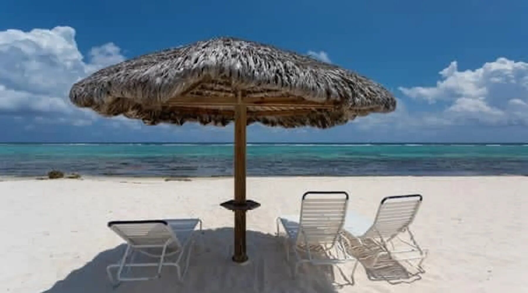 A beach with a thatched umbrella and two white lounge chairs.