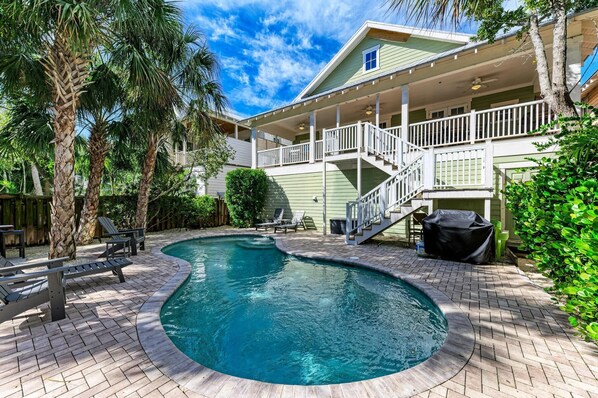 A swimming pool with a curved edge, surrounded by a brick patio. There are lounge chairs and a covered grill area.