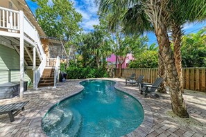 A backyard with a swimming pool, a wooden deck, a covered patio, and a fenced area with chairs.