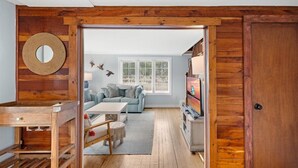 A cozy living room with a wooden wall, a round mirror, a sofa, a coffee table, and a TV. - Beachgrass Bungalow by Seaside Vacations & Sales (Chincoteague Island)