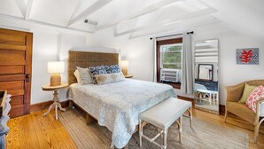 A bedroom with a bed, bedside tables, a bench, a wicker chair, a window with curtains, and a view of the outdoors. - Beachgrass Bungalow by Seaside Vacations & Sales (Chincoteague Island)