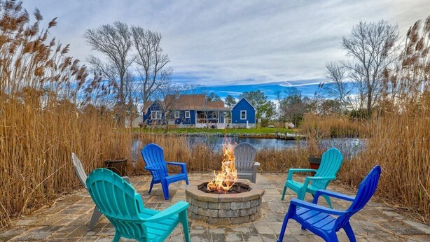 A fire pit with chairs around it, a pond, and a house in the background. - Beachgrass Bungalow by Seaside Vacations & Sales (Chincoteague Island)