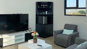 A modern living room with a flat-screen TV, a black entertainment unit with glassware, a white coffee table with a vase of flowers, and grey sofas. - HOP Encanto Exclusiva de Campo en Pisco (El Carmen)