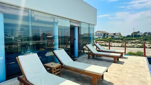 A modern building with large glass windows, a patio with lounge chairs, and a view of the sea and other buildings. - HOP Aruba Exclusiva Oasis y Playa en Chincha (El Carmen)