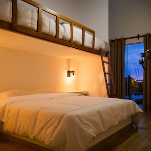 A bunk bed with a wooden frame and white bedding, a small bedside table with a light, and a view of the outside through a window with curtains.