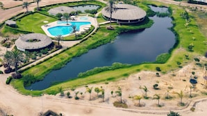 Eine Vogelperspektive zeigt einen Urlaubsort mit einem zentralen Schwimmbecken, kreisförmigen Wasserspielen und verschiedenen angelegten Grünflächen, darunter ein Spielplatz und ein kleines Gebäude.