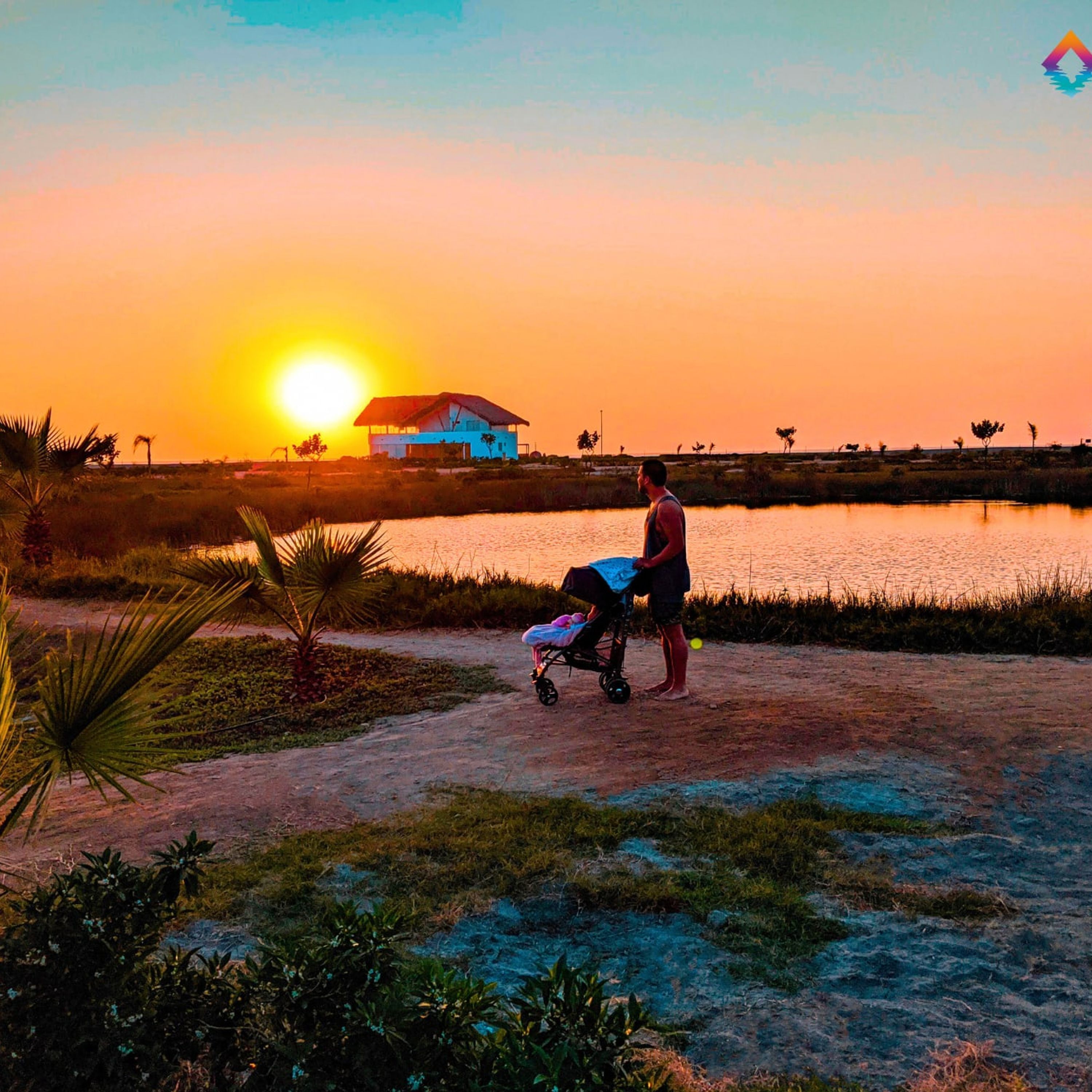 A sunset scene with a house and a person pushing a stroller by a body of water.