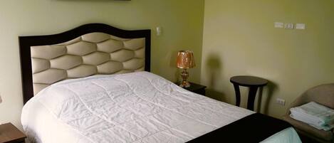 A neatly made bed with a white quilt, a headboard with a geometric pattern, a bedside lamp, a small round table, and a chair with a folded blanket.