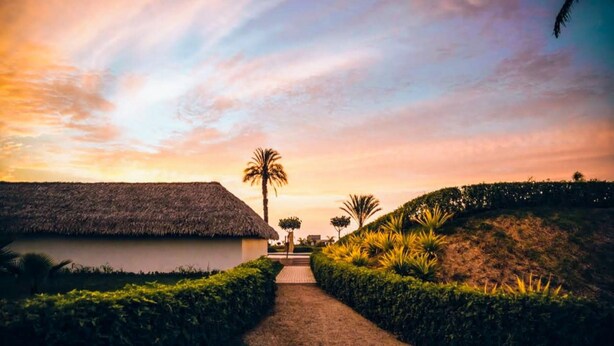 A pathway leading to a thatched-roof building, surrounded by greenery and palm trees, under a sunset sky. - HOP Odaiba Exclusiva Laguna y Playa en Chincha (El Carmen)
