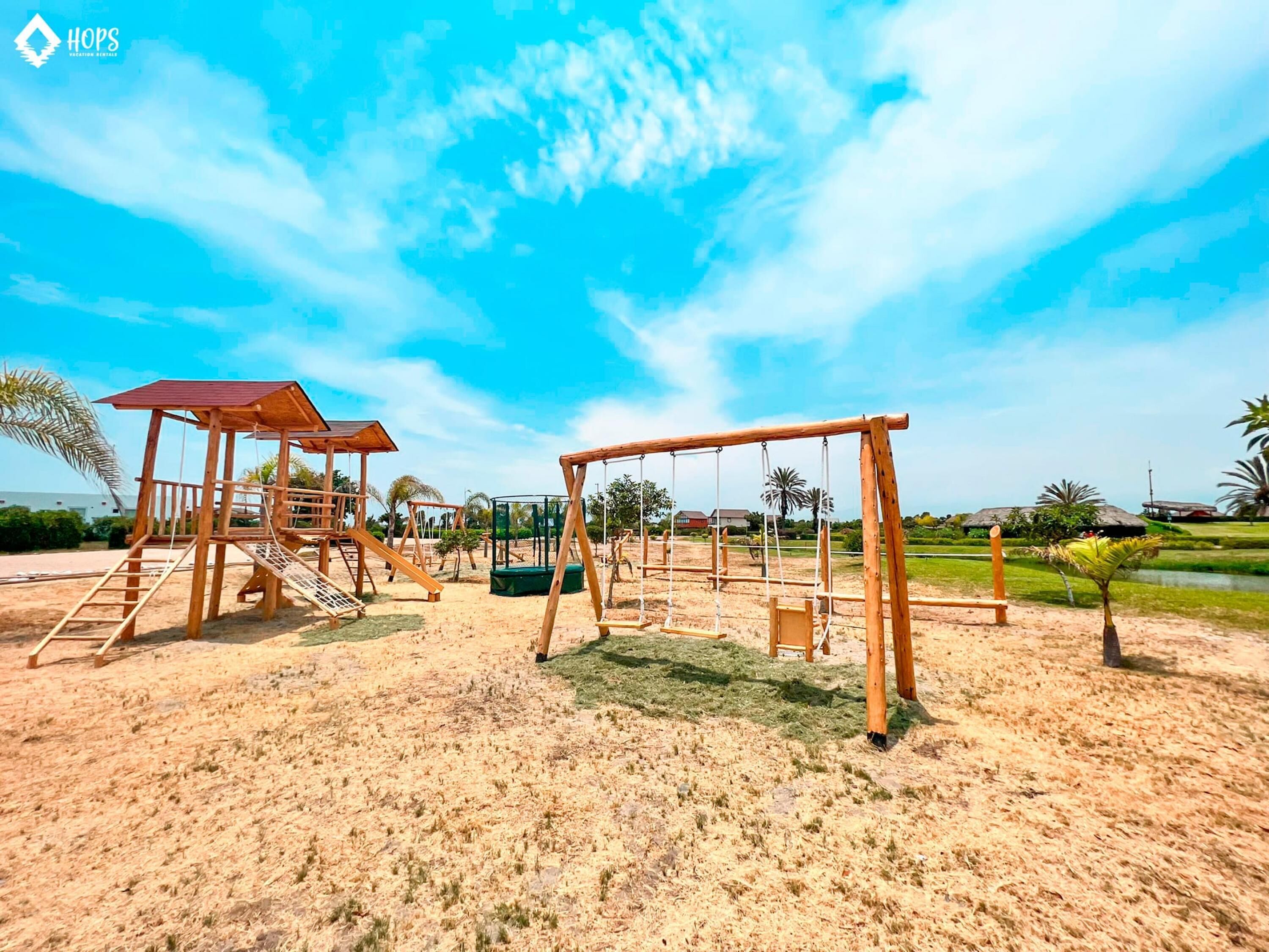 A playground with wooden structures, including swings and climbing frames, set in a sandy area with palm trees and houses in the background.