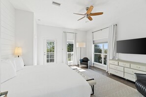 A spacious bedroom with a large bed, a ceiling fan, a TV mounted on the wall, and a view of the outdoors through the windows. - Beach House - The Sandcastle by PHG (Panama City Beach)