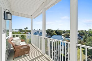 A covered balcony with a wicker chair and cushions, white railings, and a view of a blue-roofed building and surrounding greenery. - Beach House - The Sandcastle by PHG (Panama City Beach)