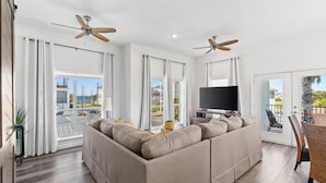 A modern living room with a sectional sofa, a flat-screen TV, ceiling fans, and large windows offering a view of the outdoors. - Beach House - The Sandcastle by PHG (Panama City Beach)