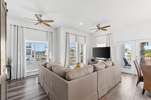 A modern living room with a sectional sofa, a flat-screen TV, ceiling fans, and large windows offering a view of the outdoors. - Beach House - The Sandcastle by PHG (Panama City Beach)