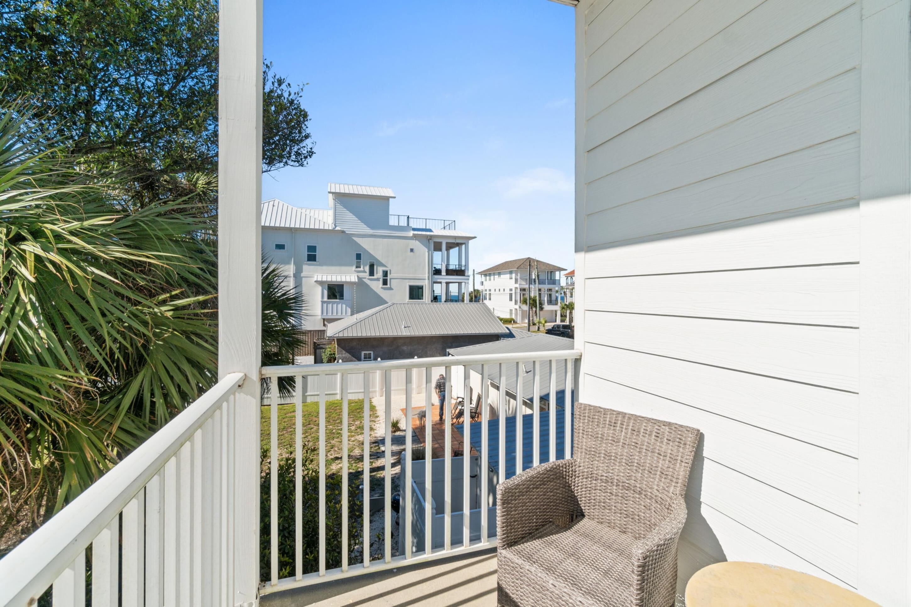 A balcony with a wicker chair and a small table, overlooking a residential area with houses and a clear sky.