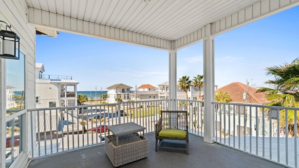 A covered balcony with a wicker chair and a small table, overlooking a residential area with houses and a clear sky. - Beach House - The Sandcastle by PHG (Panama City Beach)