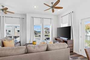 A living room with a sofa, TV, ceiling fans, and large windows with curtains. - Beach House - The Sandcastle by PHG (Panama City Beach)