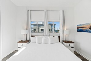 A hotel room with a large bed, two bedside tables with lamps, a view of a building through the window, and a painting on the wall. - Beach House - The Sandcastle by PHG (Panama City Beach)