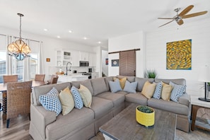 A modern living room with a sectional sofa, a coffee table, and a dining area with wicker chairs. - Beach House - The Sandcastle by PHG (Panama City Beach)