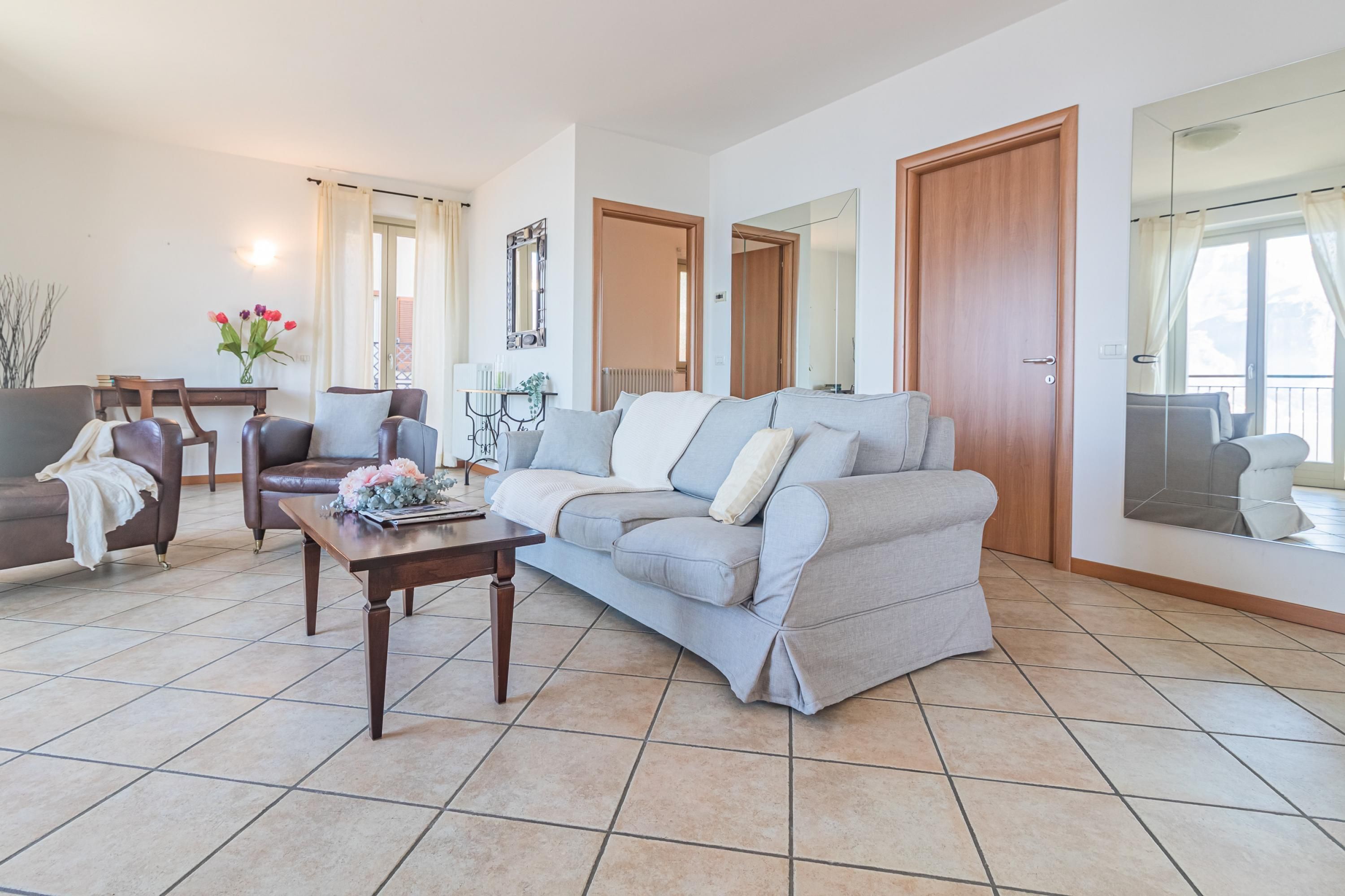 A living room with a sofa, armchairs, a coffee table, and a dining table. There are doors leading to other rooms and a large window with curtains.