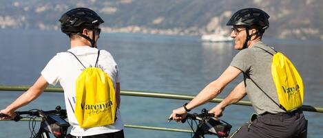 Zwei Radfahrer mit gelben Taschen, einer fährt ein Fahrrad, der andere steht daneben, vor einer Geländer und einem See mit Bergen im Hintergrund.