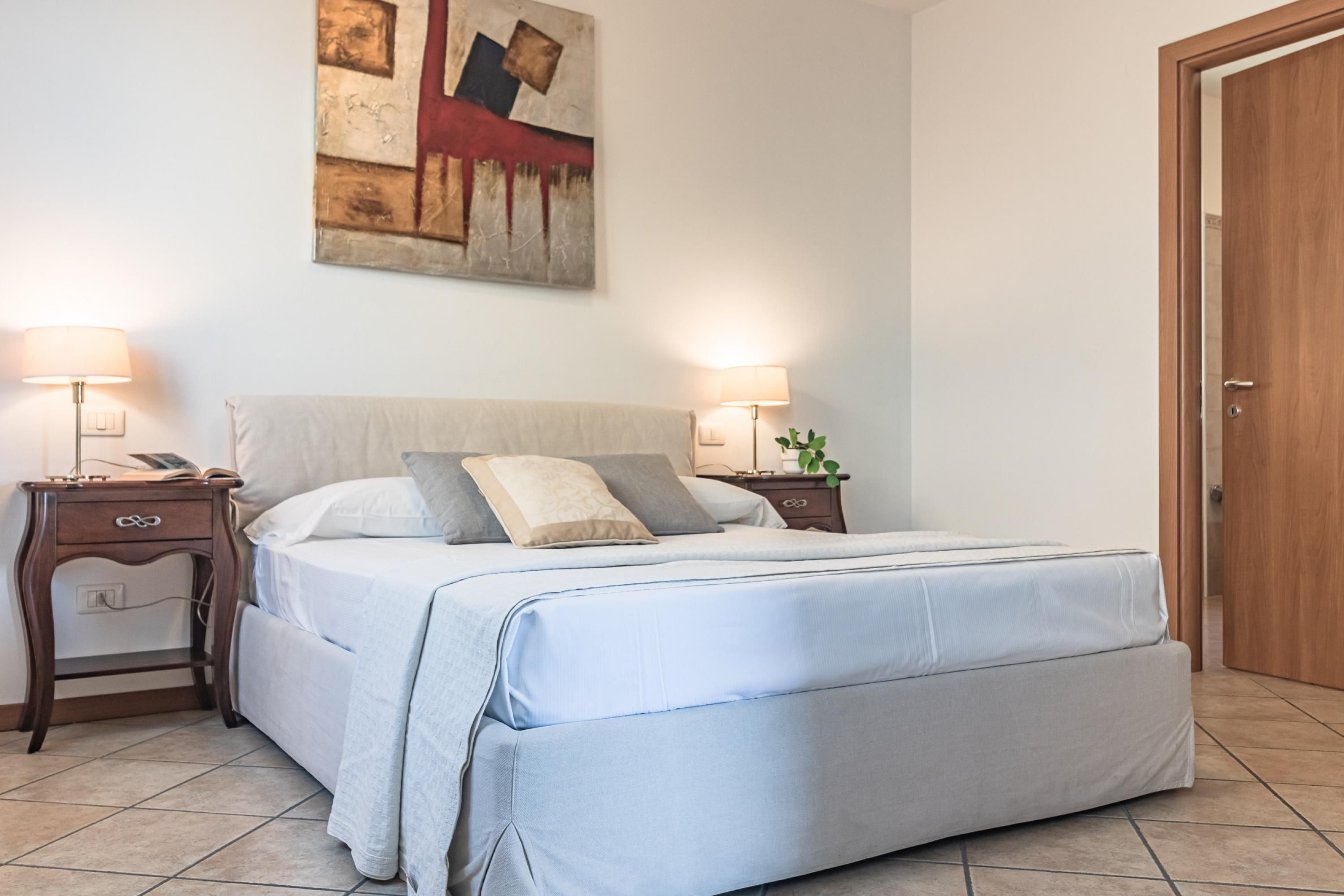 A neatly made bed with a light blue bedspread and pillows, flanked by two bedside tables with lamps and a small plant.